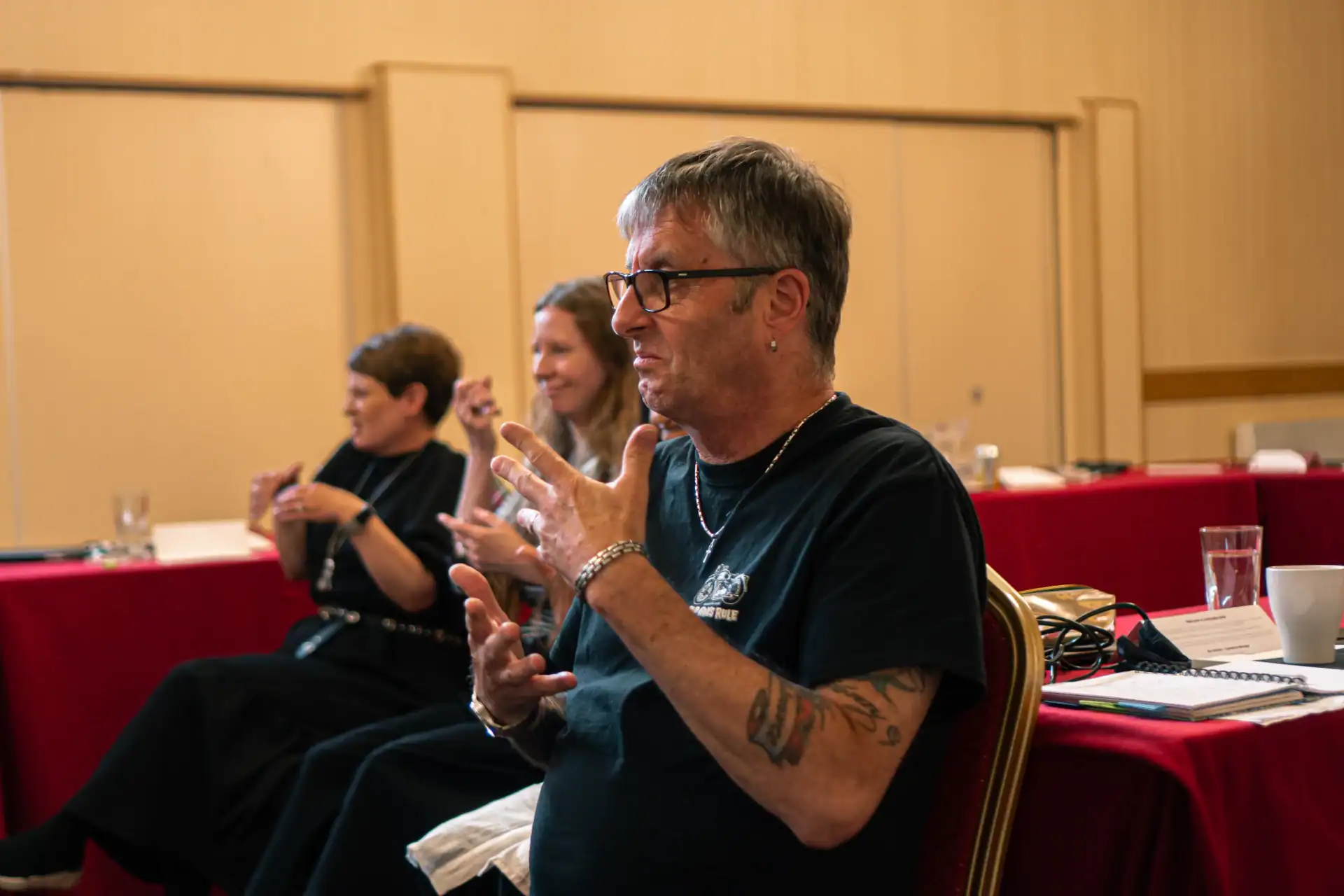 A person with tattoos and glasses uses sign language during a meeting, with two others seated in the background at a table covered in red cloth.