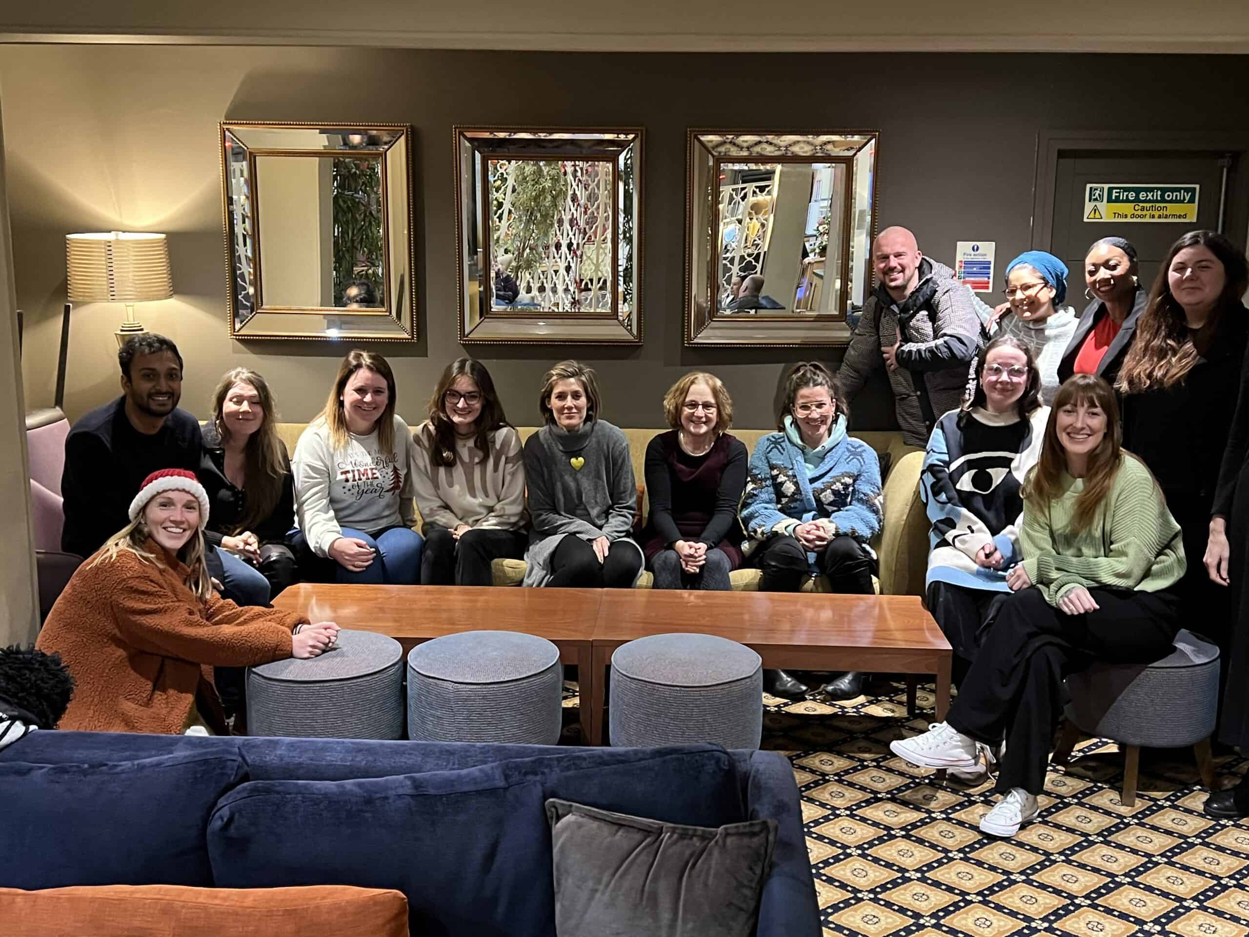 A group of fifteen people sit and stand together in a lounge area with sofas, a wooden table, and wall mirrors in the background.