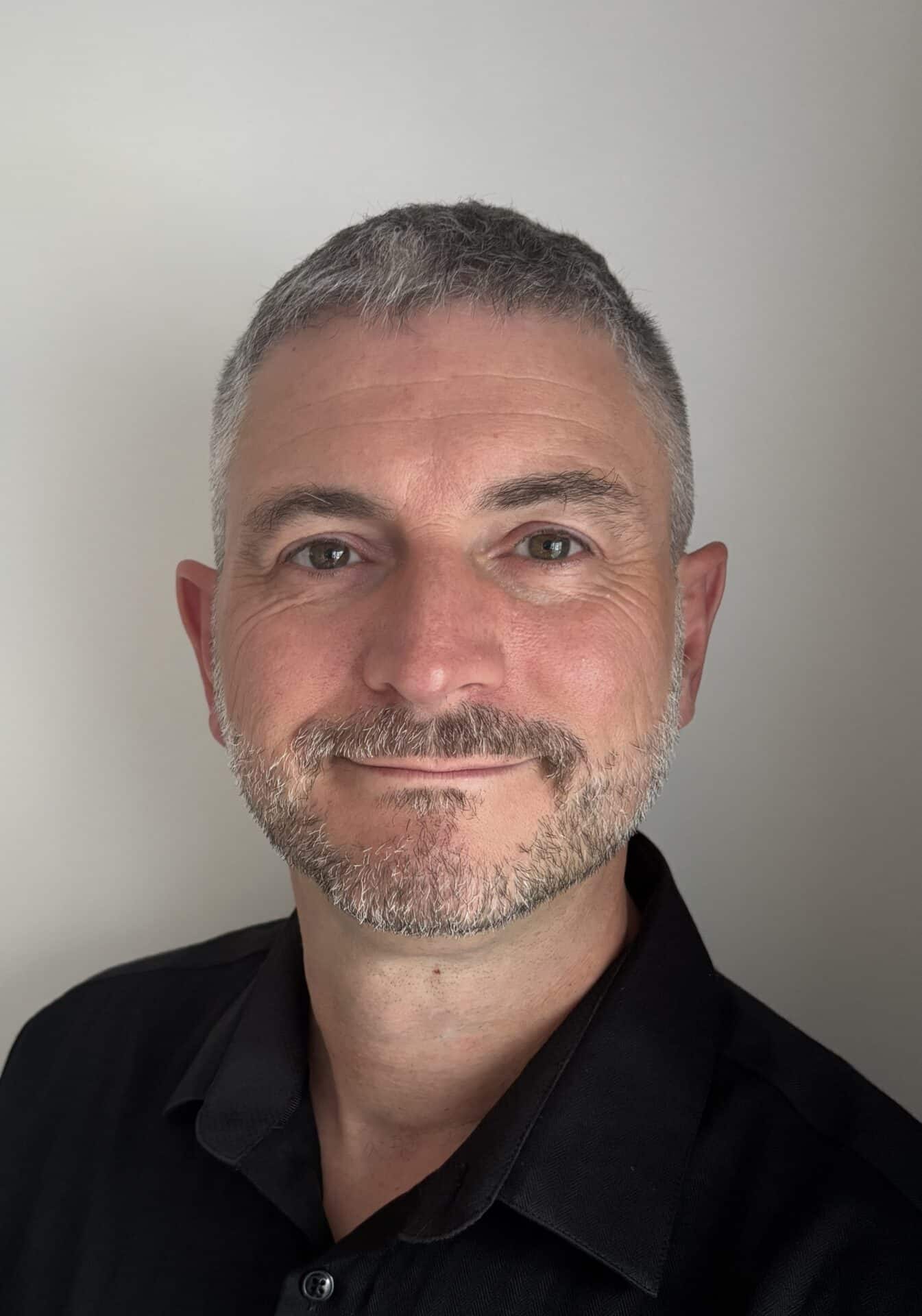 A man with short gray hair and a trimmed beard, wearing a black collared shirt, smiles slightly in front of a plain light background.