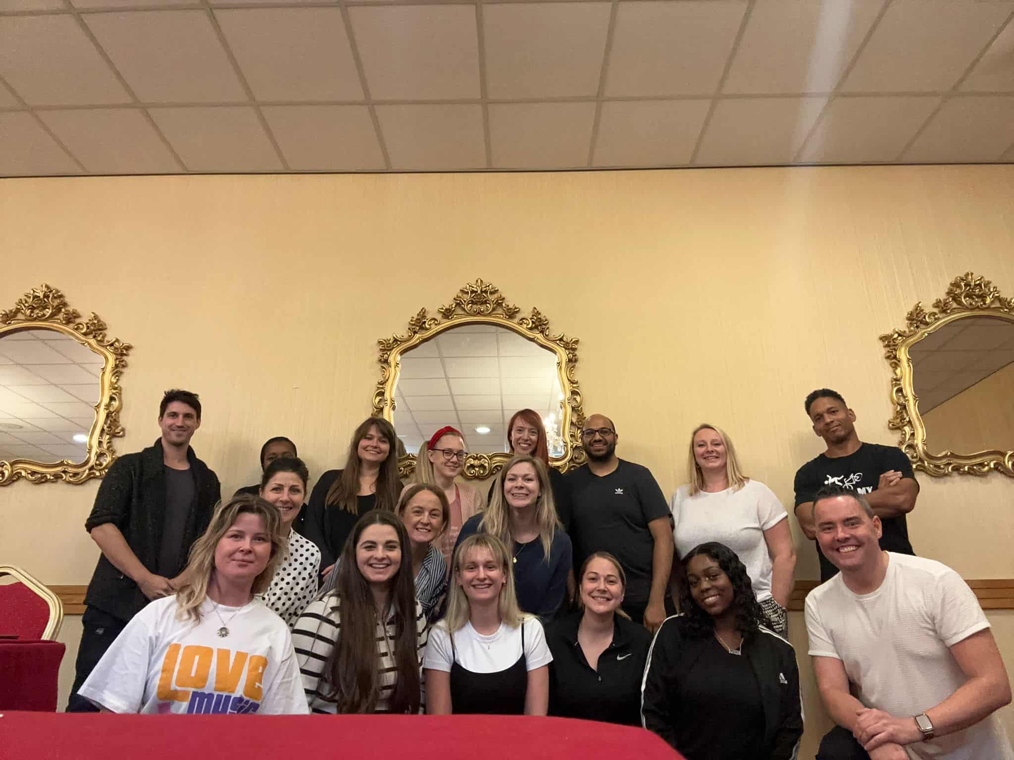 A group of sixteen people pose together indoors in front of three ornate mirrors on a cream-colored wall.