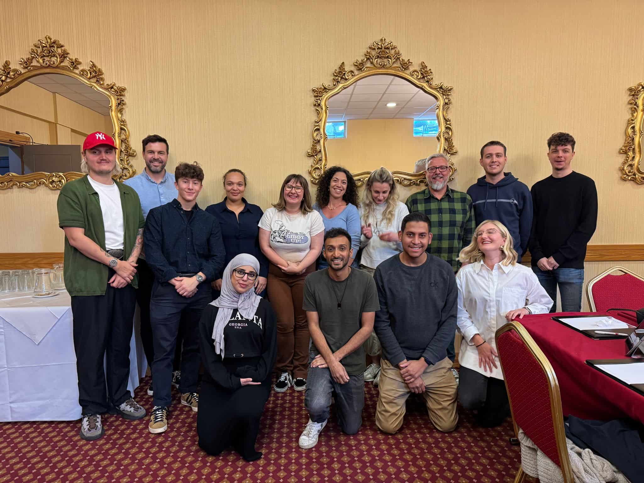 A group of fifteen people pose for a photo in a room with ornate mirrors, red carpet, and beige walls. Some are standing, others kneeling, and there are tables and chairs nearby.