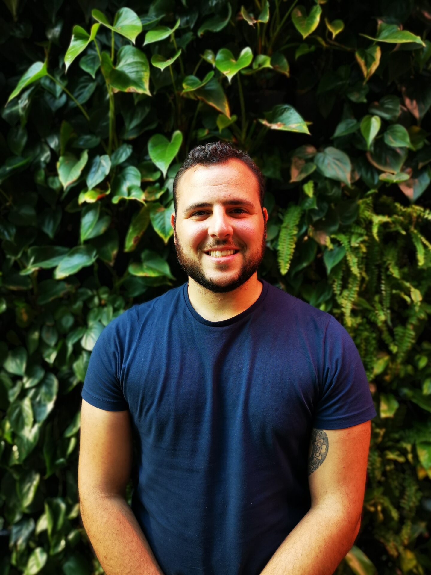 A man with short hair and a beard, wearing a navy t-shirt, stands smiling in front of a background of green leafy plants.