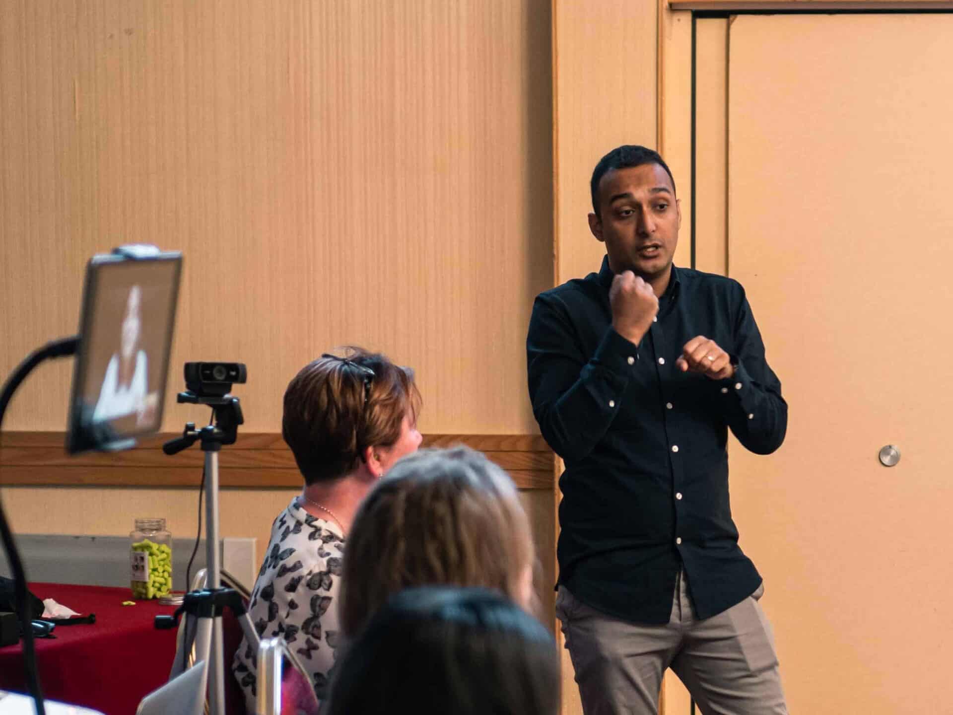 A man speaks and gestures in front of an audience, while a woman on a screen participates via video call.